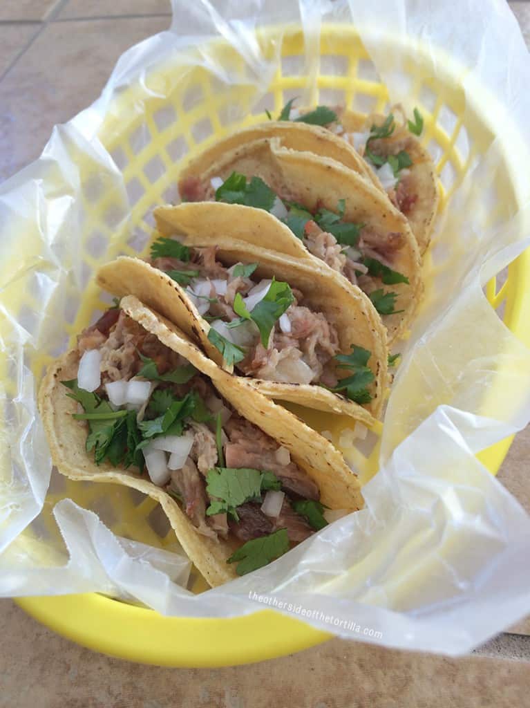 Mexican Carnitas tacos in a yellow plastic serving basket, topped with onion and chopped cilantro