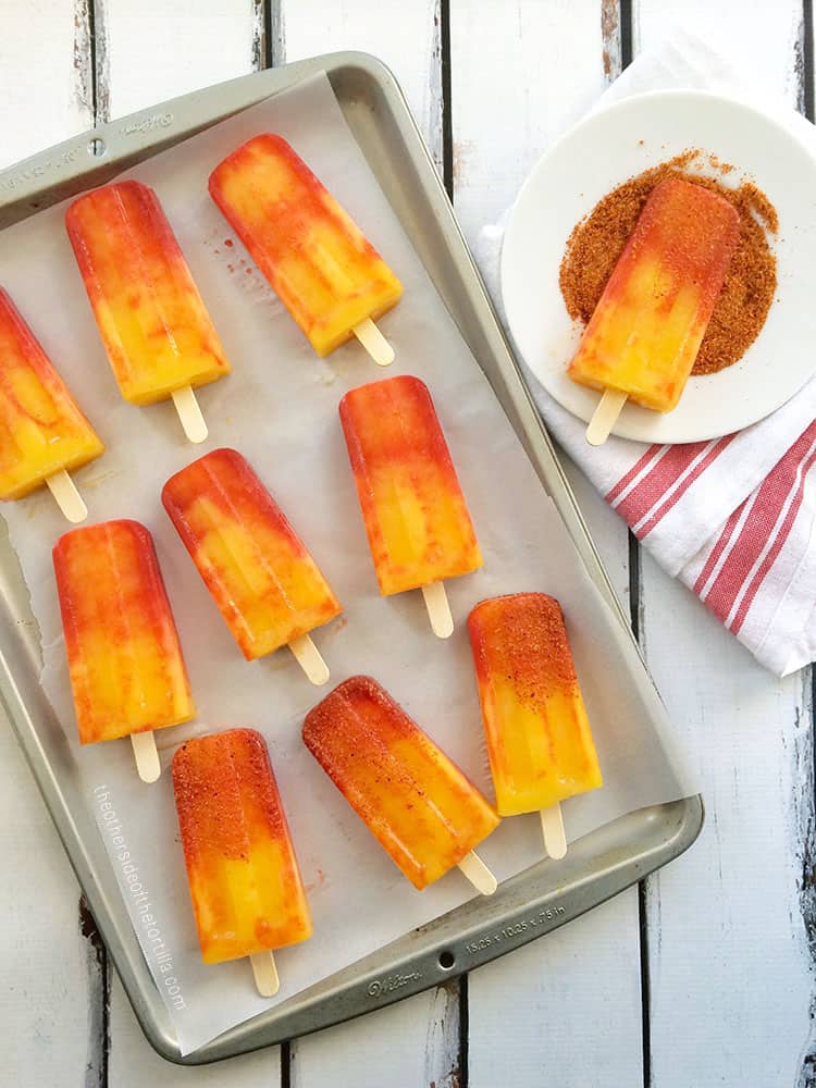 mangonada popsicles displayed on a baking sheet and a plate with tajín chili lime salt for dipping