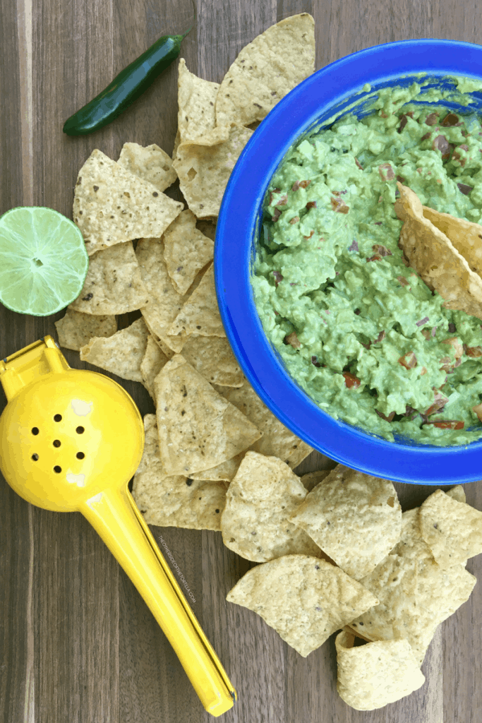 guacamole in a blue fiestaware bowl, tortilla chips, a lime and a citrus squeezer on a wood cutting board