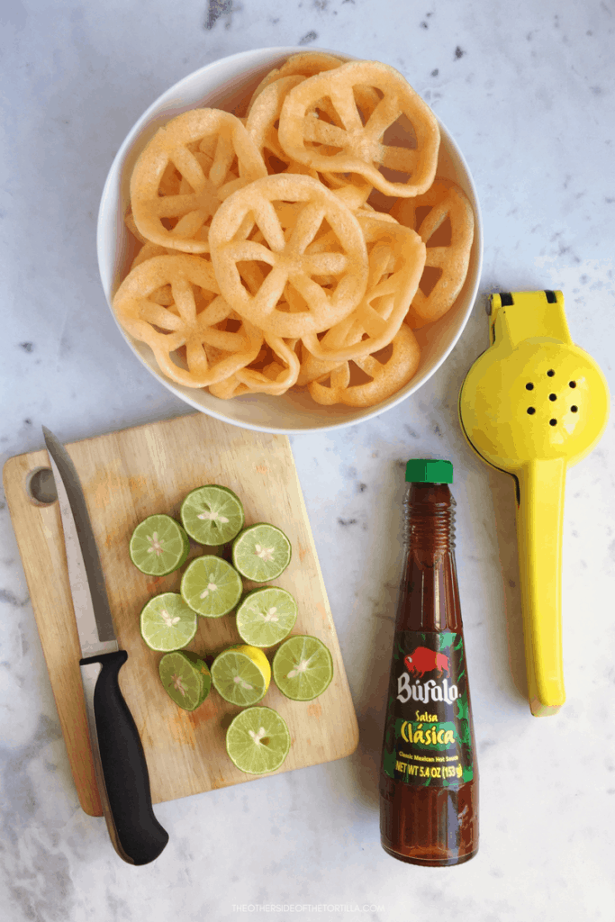 chicharrones de harina in a bowl surrounded by fresh limes on a cutting board, salsa bufalo and a lime squeezer