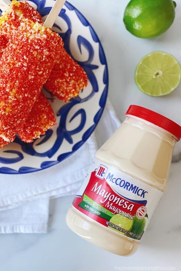 Mexican elotes locos on a blue and white talavera plate on top of a white kitchen towel surrounded by a mayonnaise jar and limes