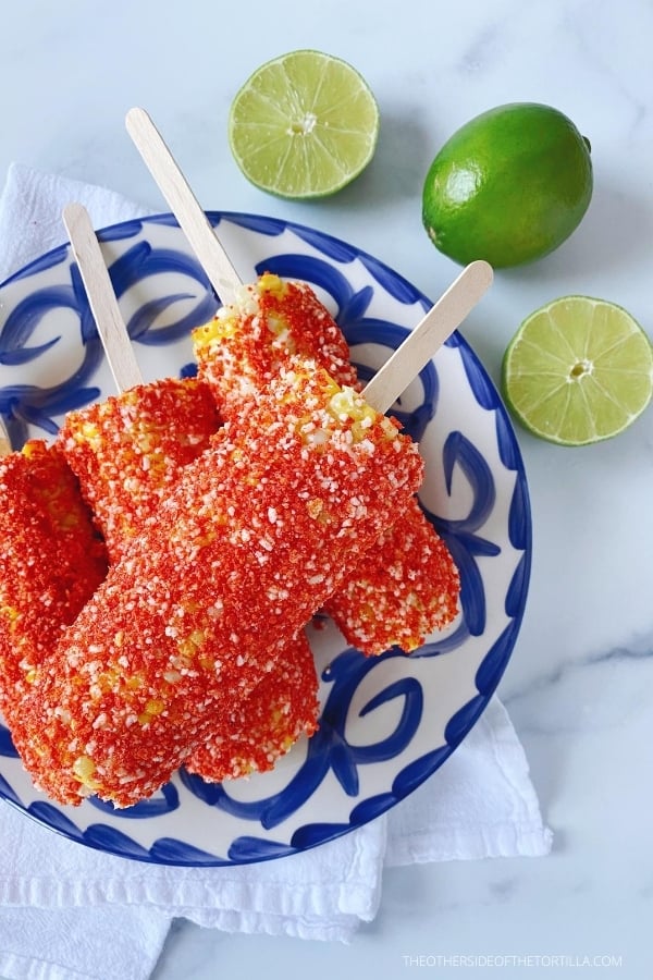 corn cobs covered in butter, mayonnaise, crushed spicy corn chips and cotija cheese, on a blue and white talavera plate, surrounded by limes