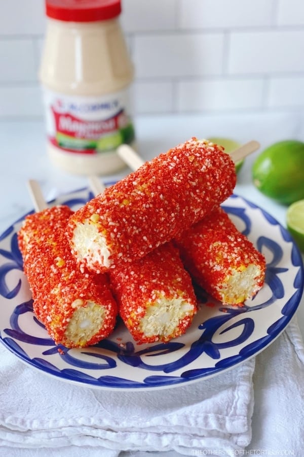 Corn cobs covered in McCormick Mayonesa with lime juice, spicy corn chip crumbs and cotija cheese, sitting on a blue and white talavera plate on top of a white dish towel and surrounded by a mayonnaise jar and limes