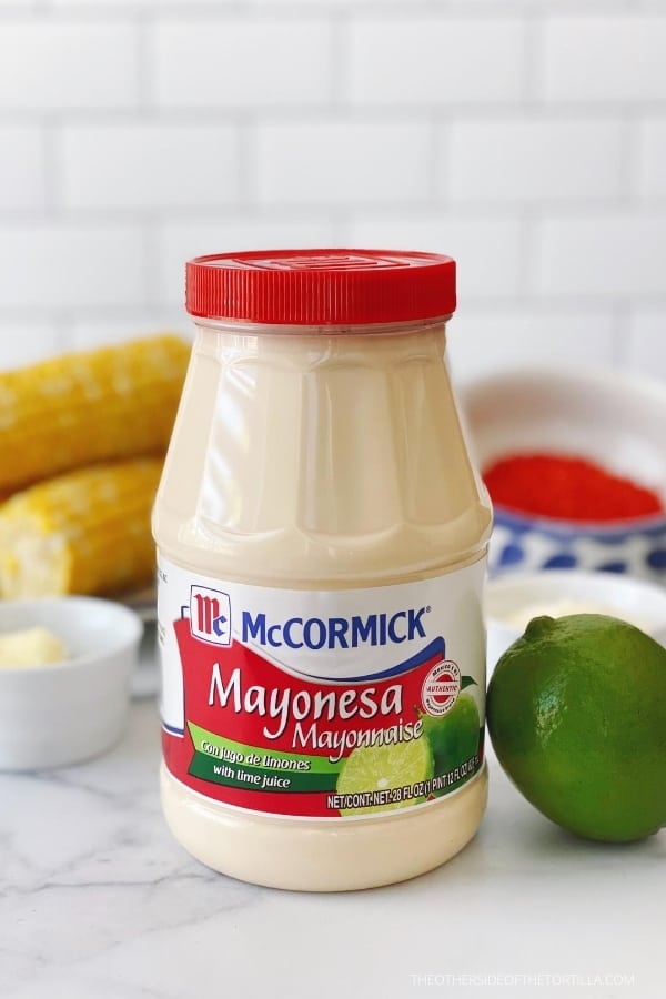 McCormick Mayonesa with lime juice on a marble countertop with a lime leaning against the jar and corn cobs in the background