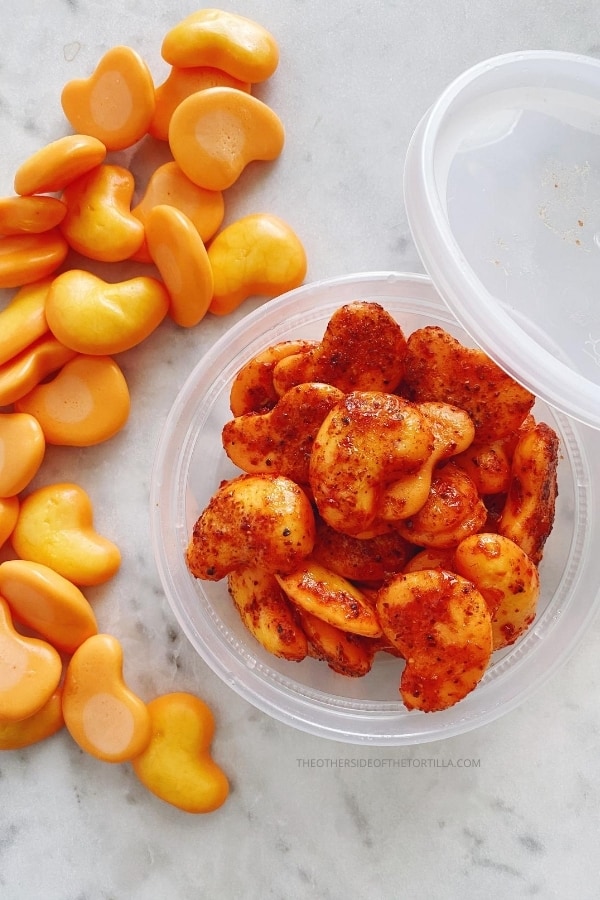 orange mango gummmies arranged on a white marble surface around a plastic container filled with spicy mango gummies dressed with Tajín and chamoy