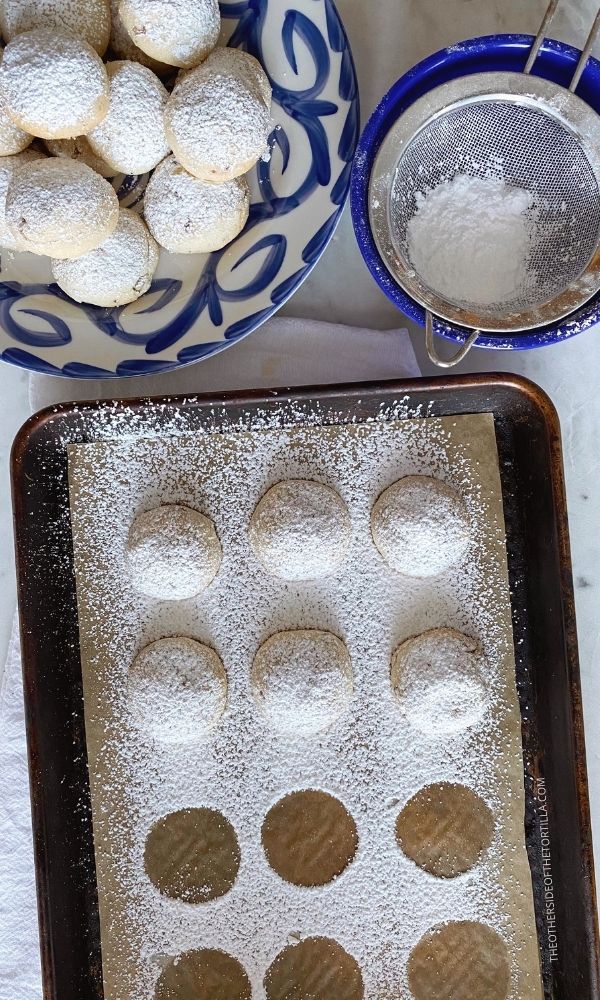 Mexican wedding cookies on a baking sheet sprinkled with powdered sugar and finished cookies on a blue and white talavera plate