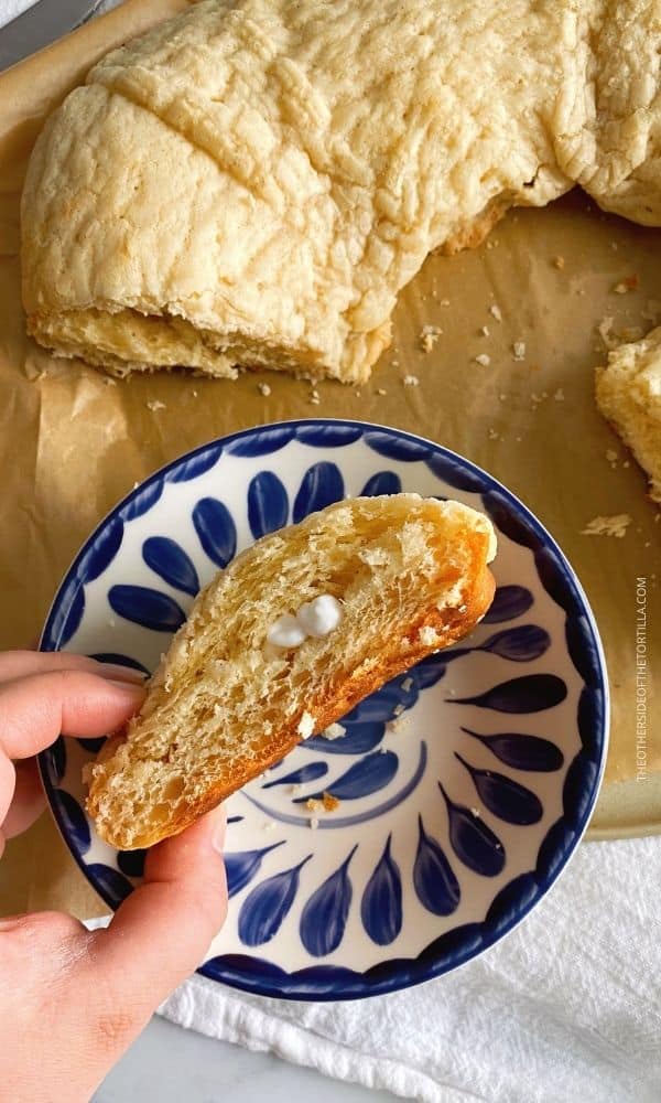 Sugar crunch rosca de reyes on a brown parchment paper at the top, handl holding a piece of sideways cake to expose the baby figurine inside, over a blue and white Mexican talavera pottery plate